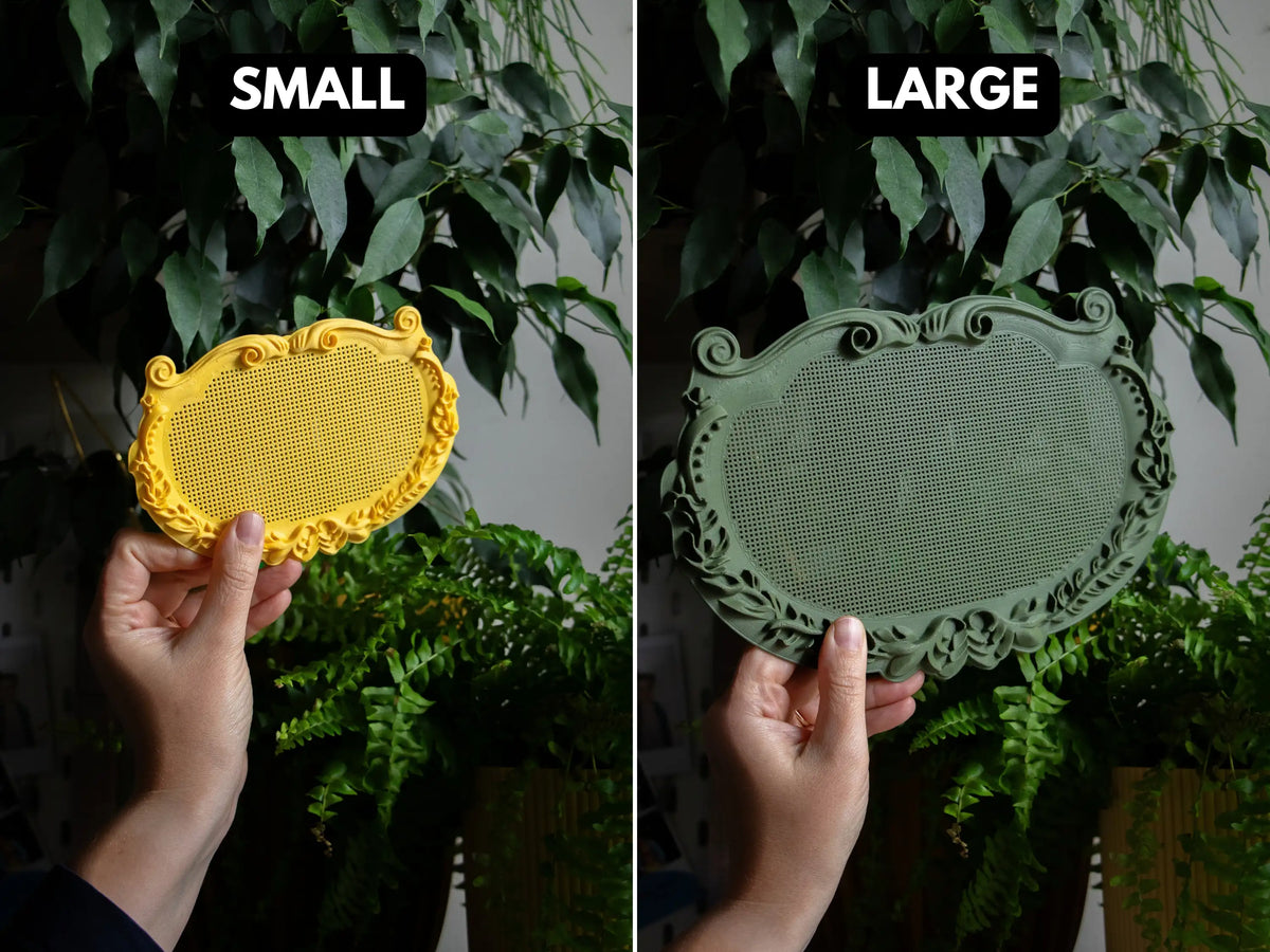 Small and large ornate plaque cross stitch frames held in hand for scale, a small yellow frame and a large sage green frame, each with a blank perforated stitching grid, in front of green houseplants.