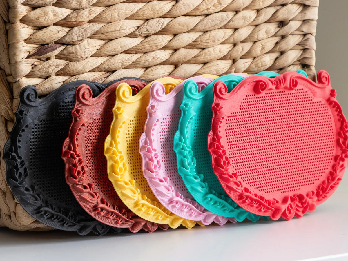 Six small ornate plaque cross stitch frames lined up in front of a woven basket, printed in black, red marble, yellow, pink, mint and red to show the design in different filament colors.