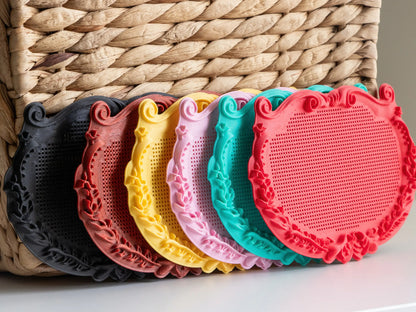 Six small ornate plaque cross stitch frames lined up in front of a woven basket, printed in black, red marble, yellow, pink, mint and red to show the design in different filament colors.
