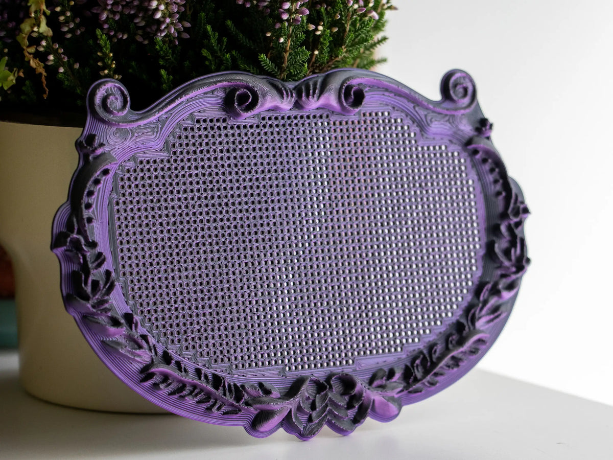 Ornate plaque cross stitch frame printed in black-purple filament, showing the scrollwork border and blank perforated stitching grid, with purple heather in the background.
