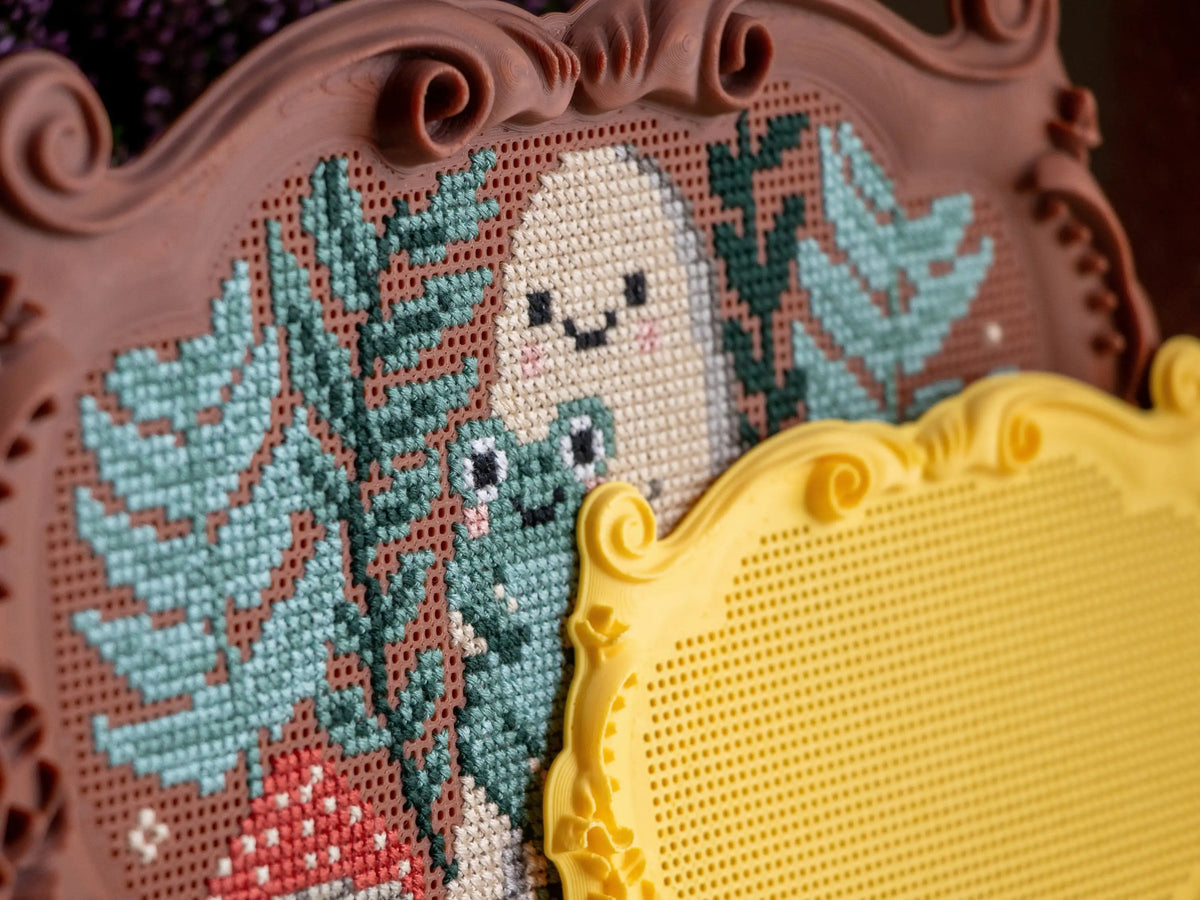 Close-up of a brown ornate plaque cross stitch frame stitched directly into the grid with a ghost, frog, leaves and a red mushroom, with a blank yellow plaque frame in front.