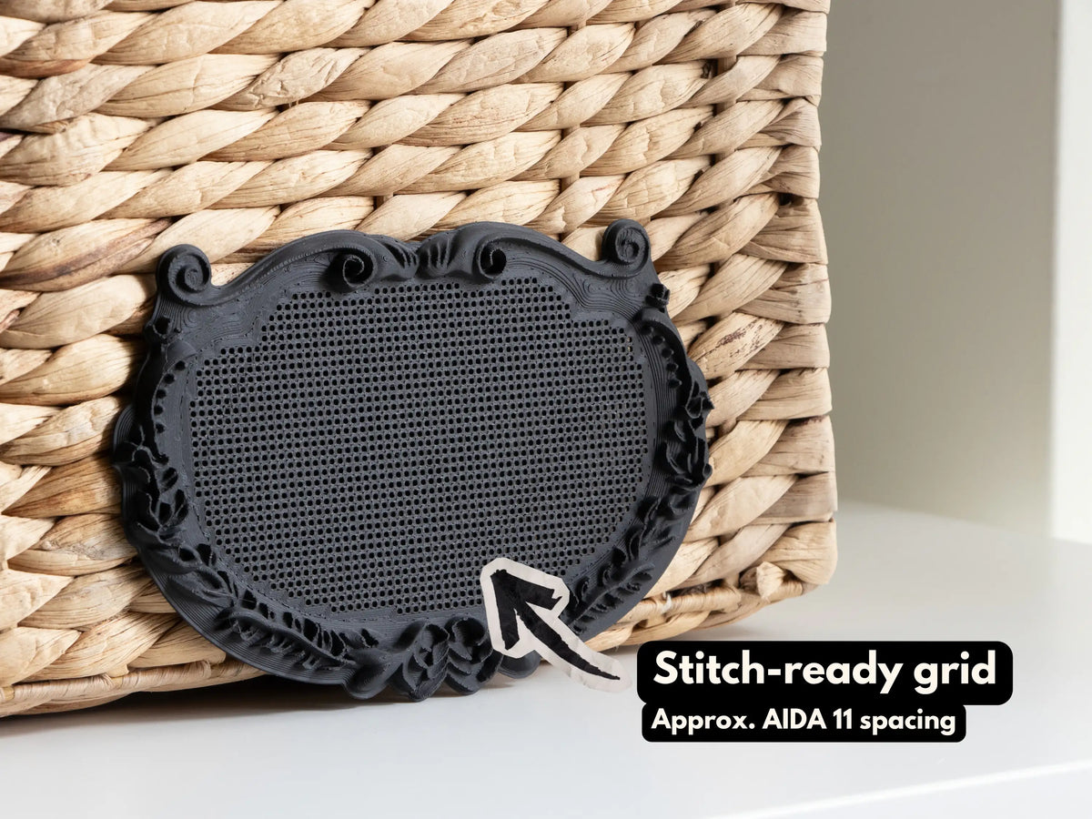 Black ornate plaque cross stitch frame leaning against a woven basket, with an arrow pointing to the blank perforated stitching grid, which has spacing close to Aida 11.