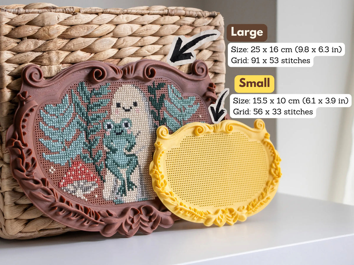 Size comparison of the ornate plaque cross stitch frames: the large 25 × 16 cm brown frame stitched with a ghost, frog and mushroom, and the small 15.5 × 10 cm blank yellow frame in front.