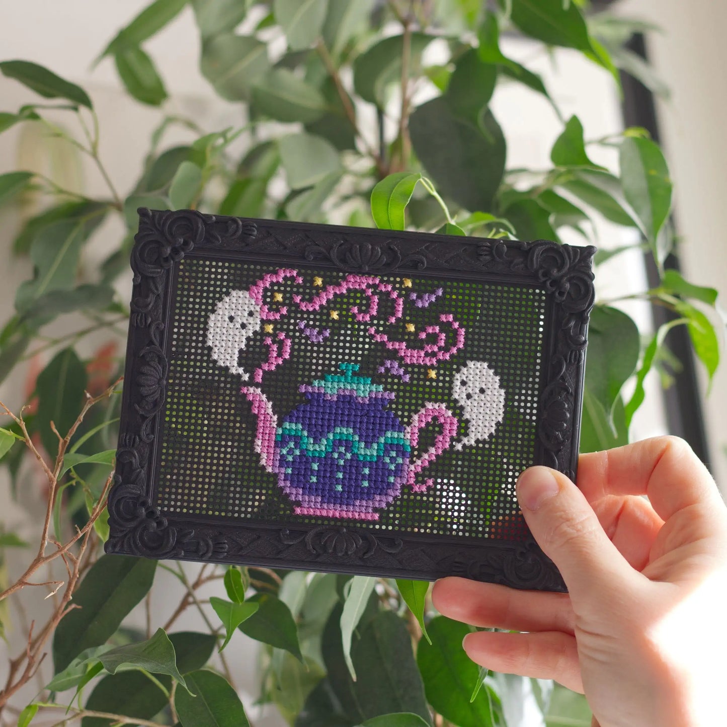 Finished cross stitch teapot design stitched directly into a decorative black stitchable embroidery frame held in hand in front of green houseplants.