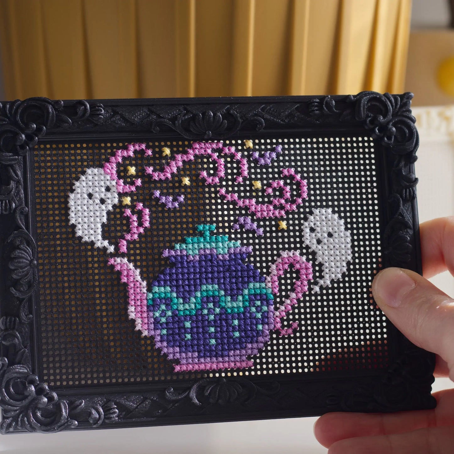 Close-up of a decorative black stitchable embroidery frame held in hand showing a finished cross stitch teapot design stitched directly into the perforated surface.