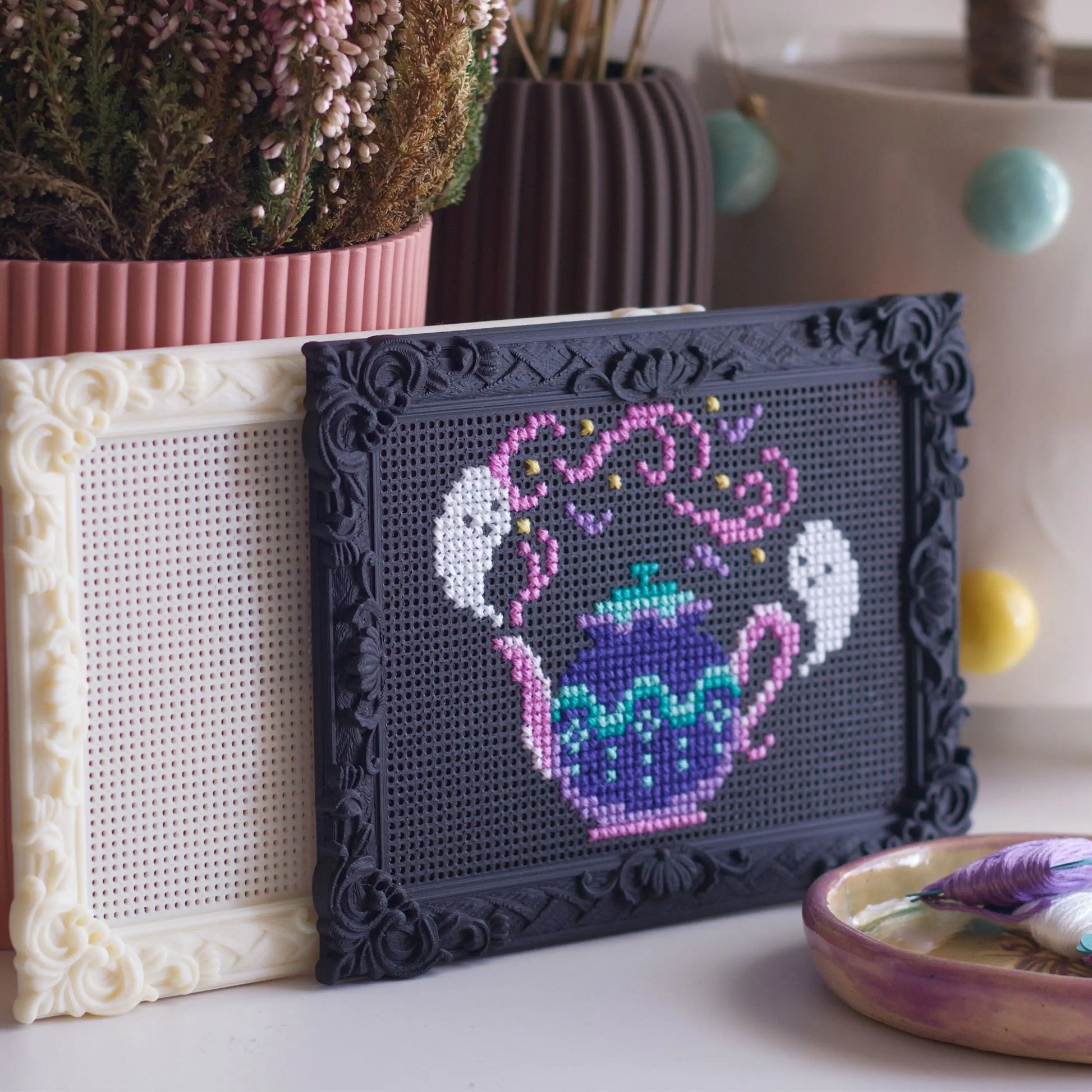 Decorative black stitchable cross stitch frame with finished teapot embroidery displayed on a shelf with a blank cream frame behind it and craft supplies nearby.