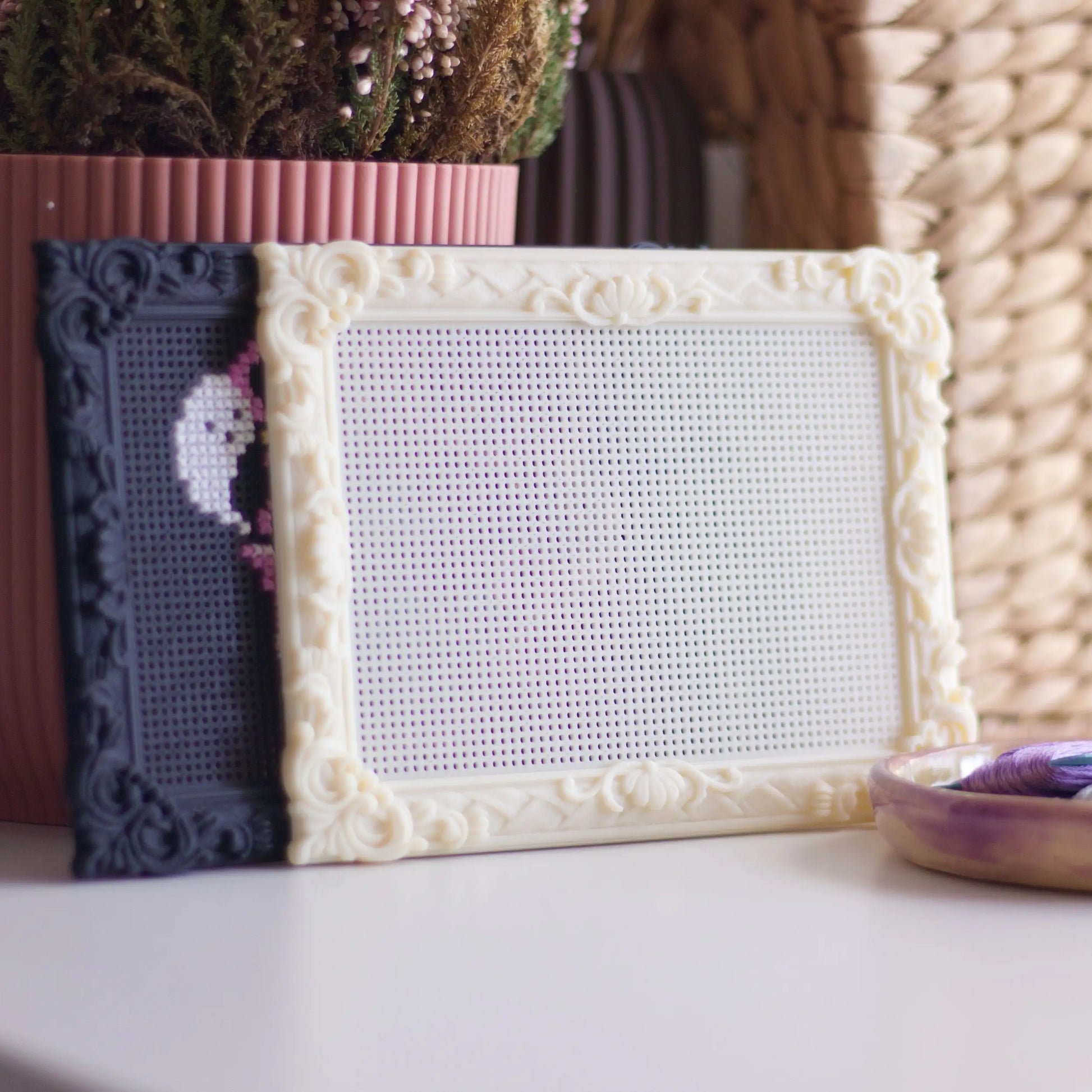 Cream and black stitchable cross stitch frames displayed on a shelf with plants and embroidery thread, showing blank perforated stitching surface.
