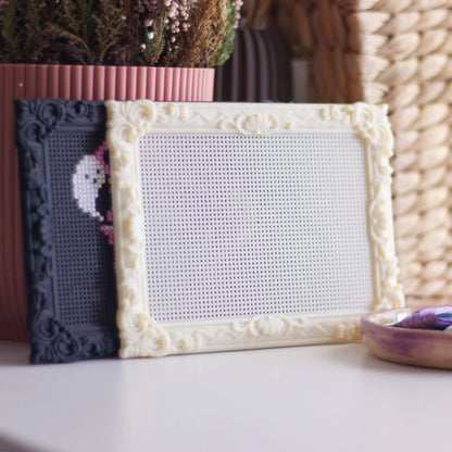 Cream and black stitchable cross stitch frames displayed on a shelf with plants and embroidery thread, showing blank perforated stitching surface.
