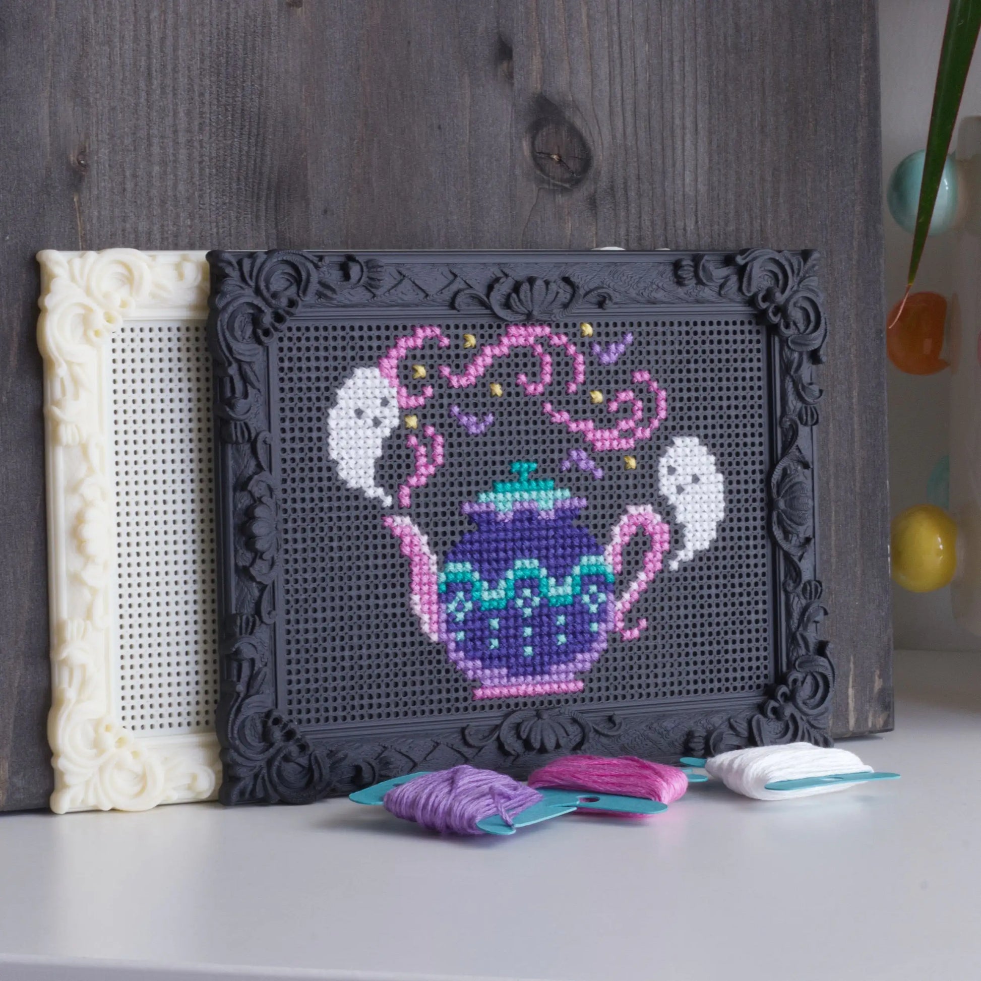 Finished cross stitch teapot design stitched into a black decorative embroidery frame with embroidery floss placed in front.
