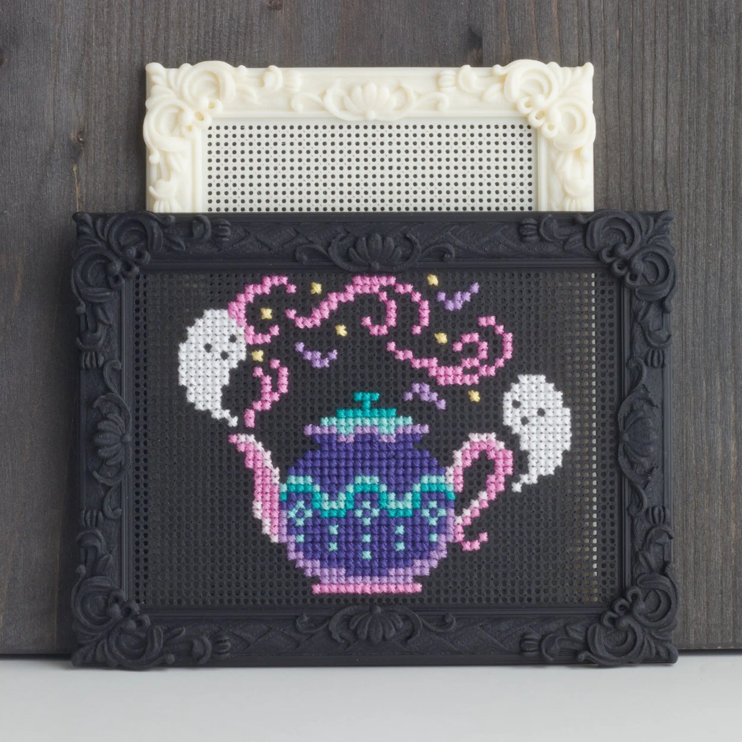 Decorative black stitchable cross stitch frame with finished teapot embroidery placed in front of a blank cream embroidery frame.