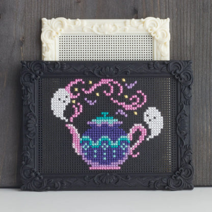 Decorative black stitchable cross stitch frame with finished teapot embroidery placed in front of a blank cream embroidery frame.