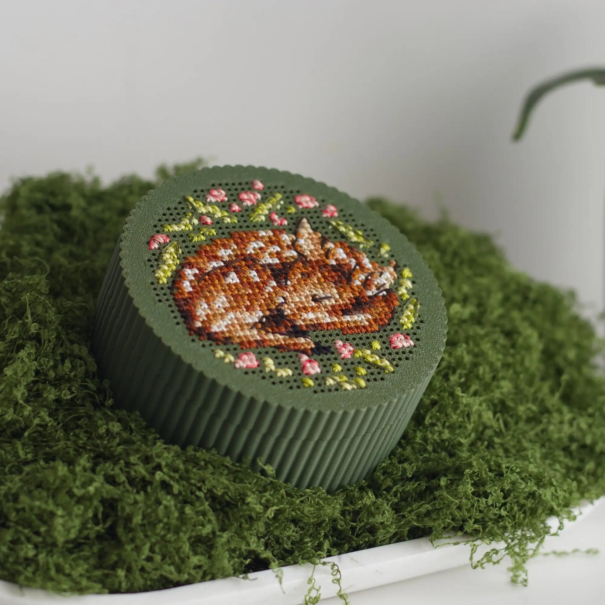 Round dark green cross stitch box with finished sleeping fawn embroidery and pink floral wreath on perforated lid, displayed on green moss tray.