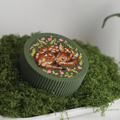 Round dark green cross stitch box with finished sleeping fawn embroidery and pink floral wreath on perforated lid, displayed on green moss tray.