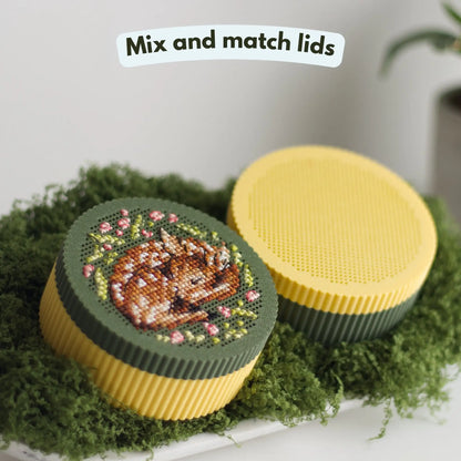 Mix and match round cross stitch boxes — yellow base with dark green stitched fawn lid beside yellow base with yellow perforated grid lid, displayed on green moss