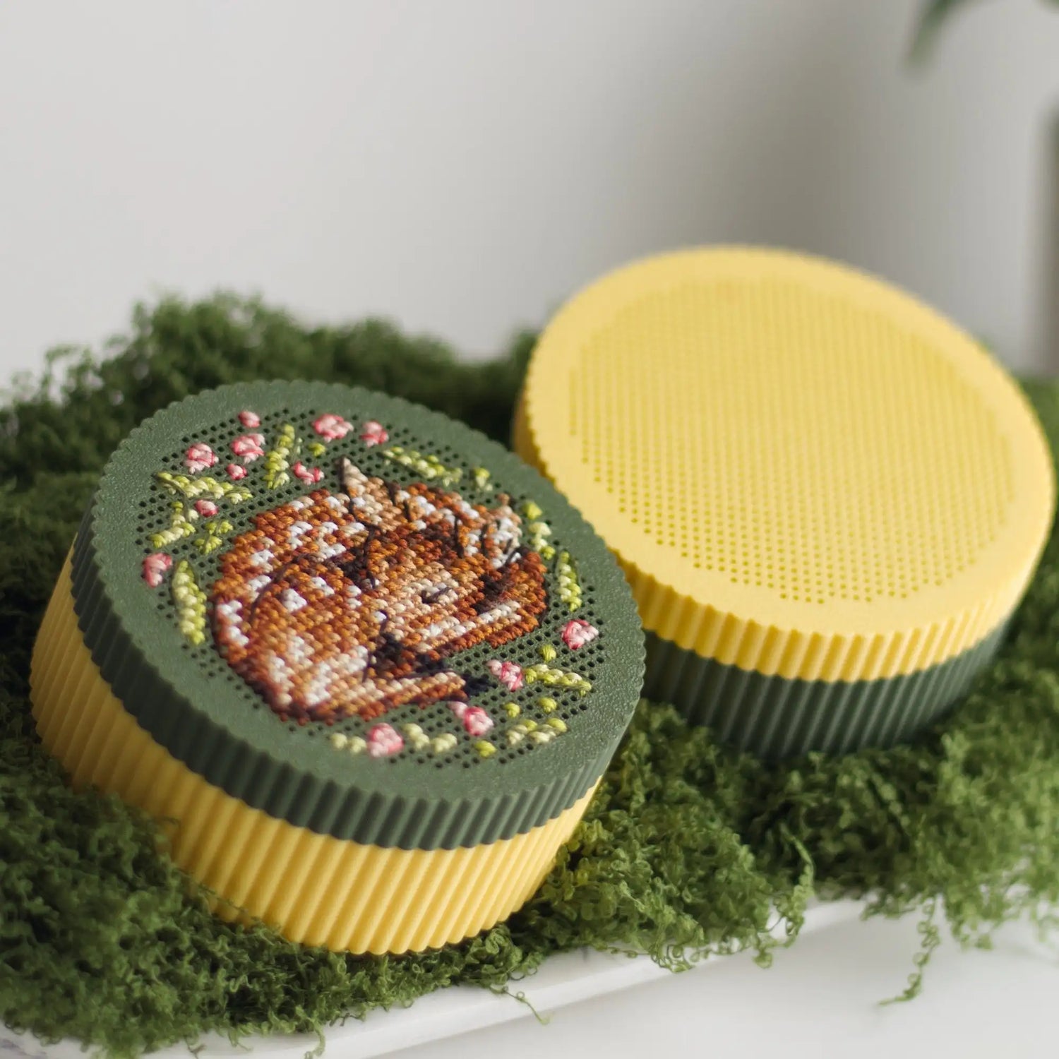 Mix and match round cross stitch boxes — yellow base with dark green stitched fawn lid beside yellow base with yellow perforated grid lid, displayed on green moss
