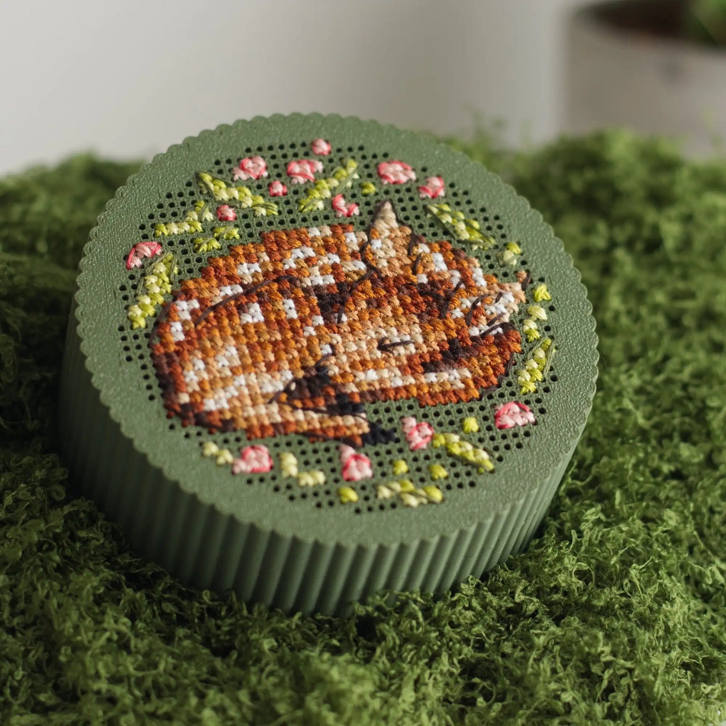 Round dark green cross stitch box with finished sleeping fawn embroidery and pink floral wreath on perforated lid, displayed on green mossRound dark green cross stitch box with finished sleeping fawn embroidery and pink floral wreath on perforated lid, displayed on green moss