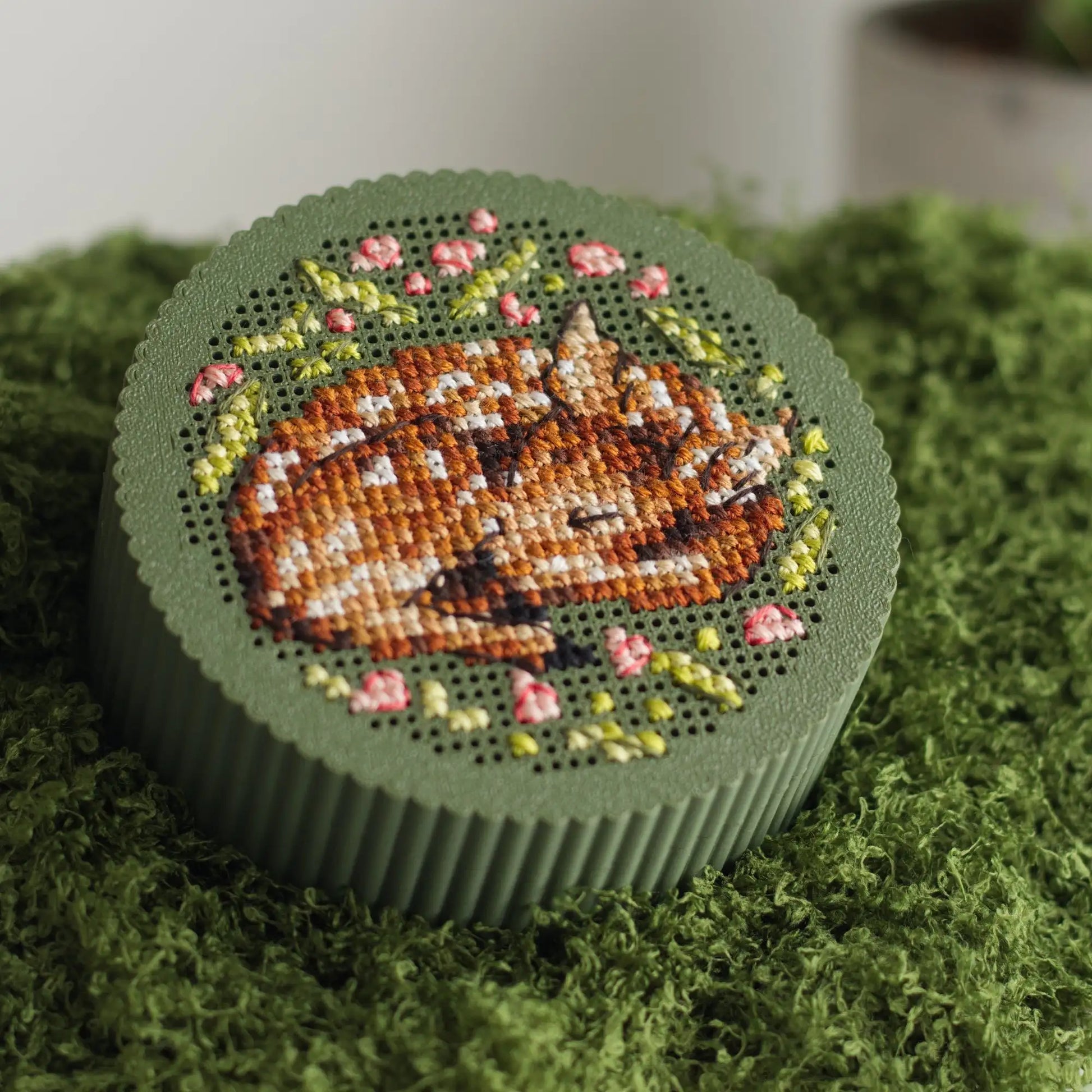 Round dark green cross stitch box with finished sleeping fawn embroidery and pink floral wreath on perforated lid, displayed on green mossRound dark green cross stitch box with finished sleeping fawn embroidery and pink floral wreath on perforated lid, displayed on green moss