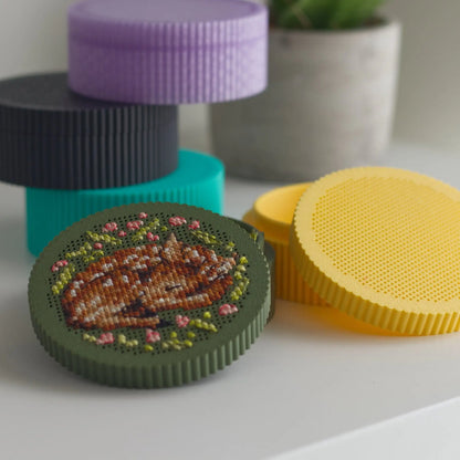 Round cross stitch boxes in dark green, yellow, teal, black, and purple — finished sleeping fawn embroidery on dark green lid in foreground