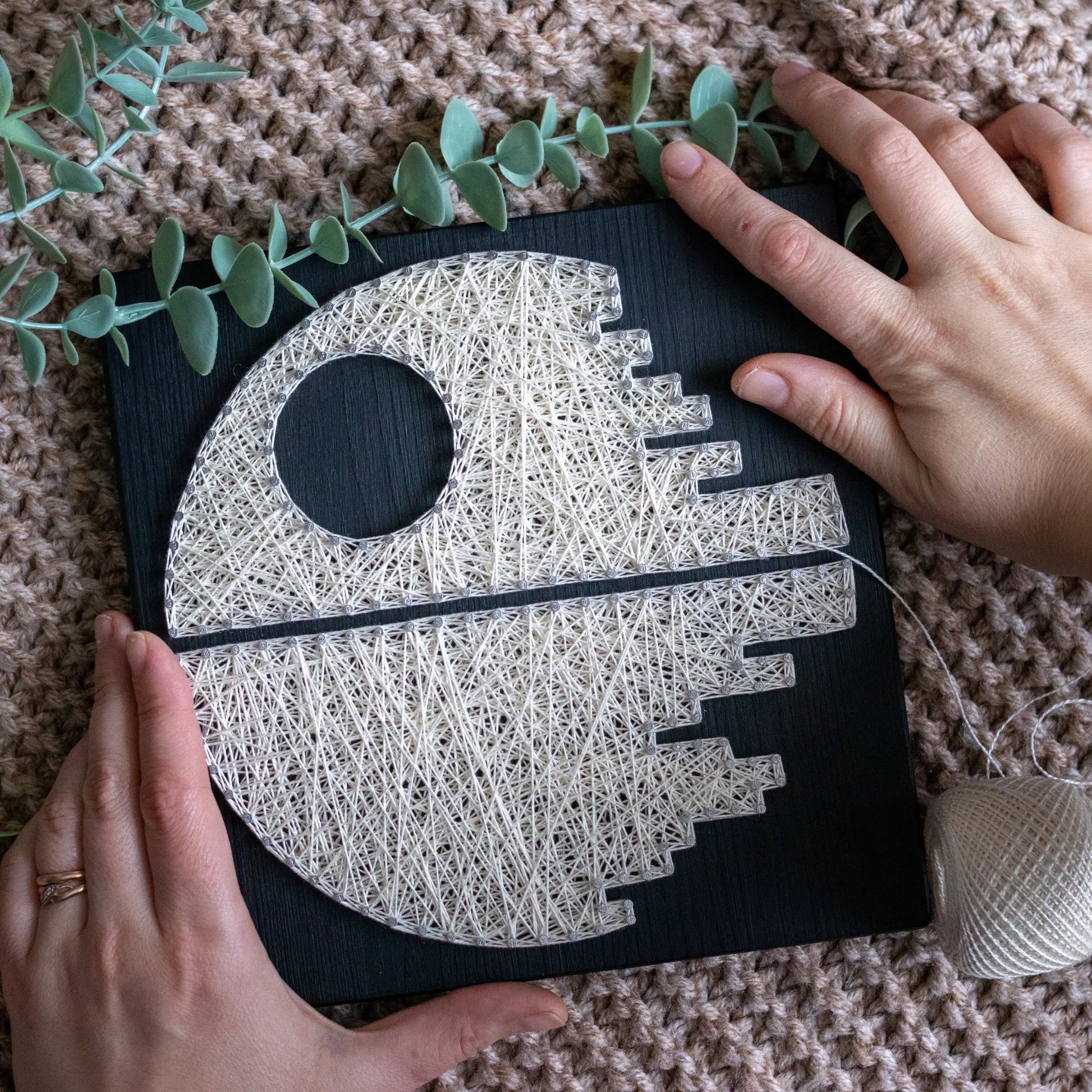 DIY string art kit featuring a Death Star design with white threads on a black wooden board by GoodStrings with top view with hands on sides