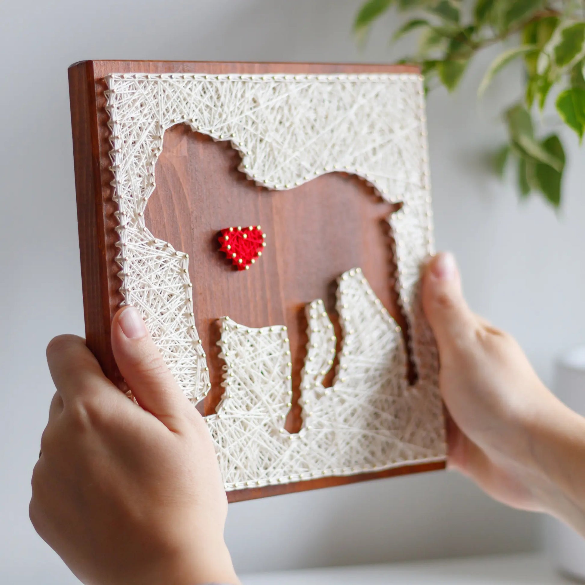 DIY string art kit featuring a dog silhouette with a red heart outlined with ivory thread on a rustic wooden board by GoodStrings - hands holding