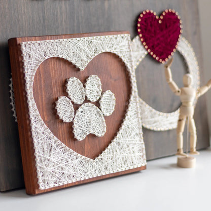 Side view. String art pattern featuring a paw print design inside a heart shape. The artwork is made using white string on a wooden board, creating a minimalist, rustic look. This digital download pattern allows you to create a similar piece at home.