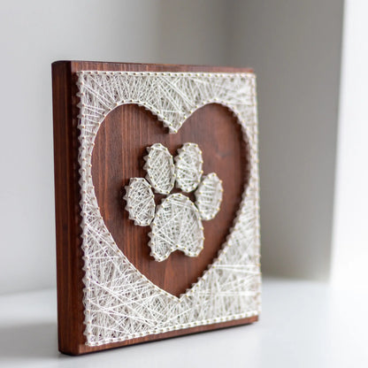 Side view of a handcrafted string art piece featuring a white string paw print inside a heart shape on a dark wooden board. The photo highlights the texture of the string and the depth of the raised design, showing the intricate details of the art.