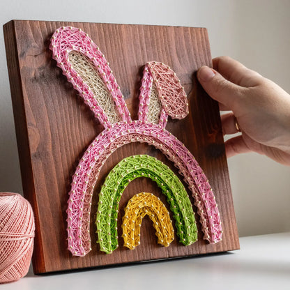 Hand holding finished Easter Bunny Rainbow string art sign made with pink, green, and yellow threads on a dark wood board.