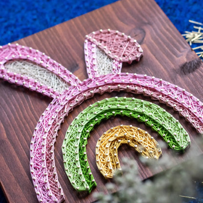 Close-up of Easter string art pattern featuring rainbow arches and bunny ears in spring colors stitched on wood.