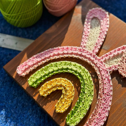 Brightly lit Easter Bunny Rainbow string art design on a wooden board, surrounded by pink and green thread spools.