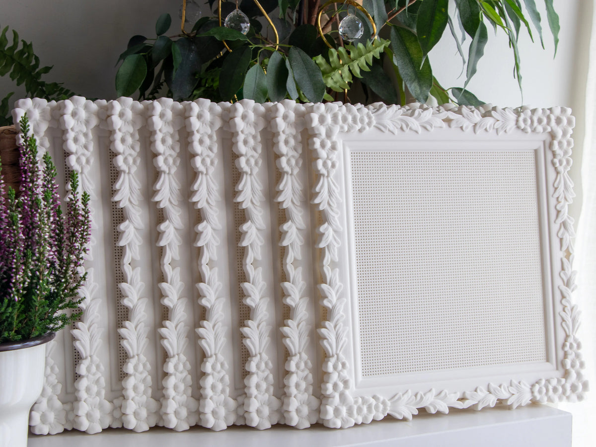 Row of white floral cross stitch frames standing upright on a shelf with one turned to face forward, showing the blank stitchable grid and the flower-and-leaf border, with heather and houseplants beside them.