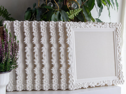 Row of white floral cross stitch frames standing upright on a shelf with one turned to face forward, showing the blank stitchable grid and the flower-and-leaf border, with heather and houseplants beside them.