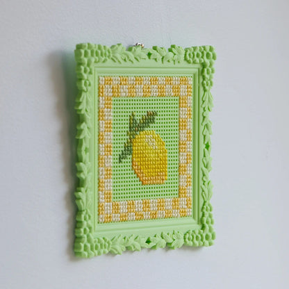 Floral cross stitch frame in green, mounted on the wall with a finished lemon cross stitch design inside.
