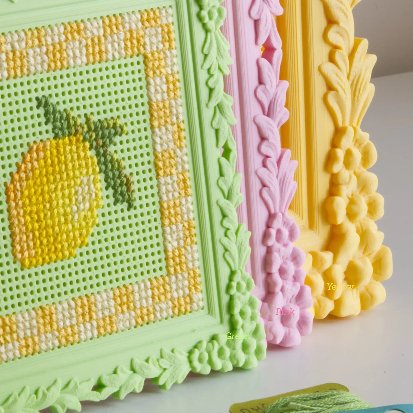 Floral cross stitch frames in green, pink, and yellow lined up, showing the finished lemon design and the ornate floral border detail up close.
