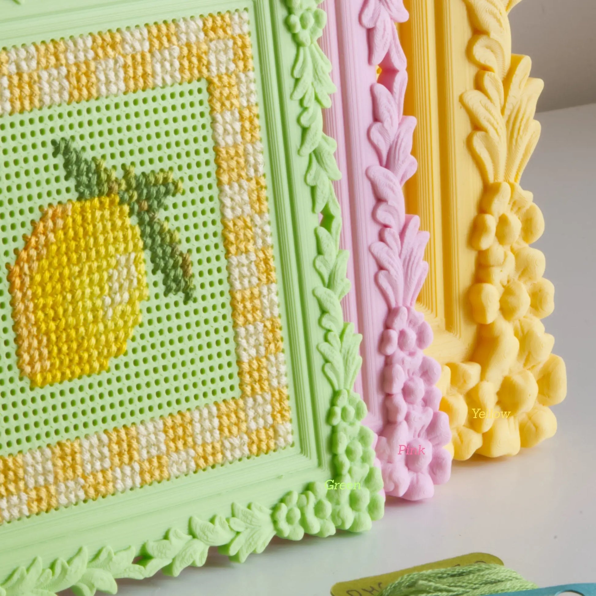 Floral cross stitch frames in green, pink, and yellow lined up, showing the finished lemon design and the ornate floral border detail up close.
