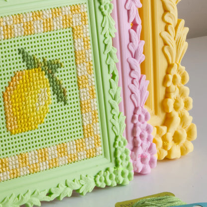 Floral cross stitch frames in green, pink, and yellow lined up, showing the finished lemon design and the ornate floral border detail up close.
