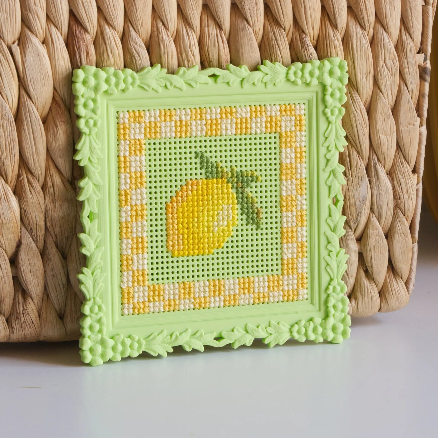 Green floral cross stitch frame with a finished lemon design, leaning against a woven basket.