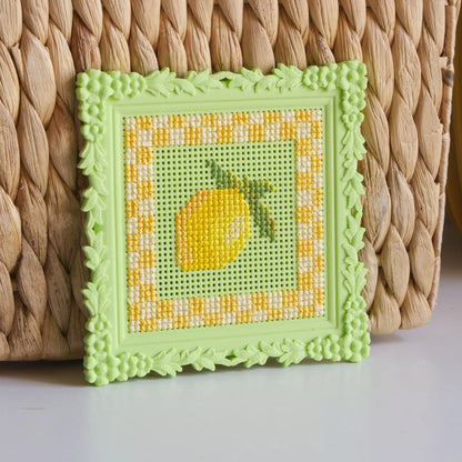Green floral cross stitch frame with a finished lemon design, leaning against a woven basket.
