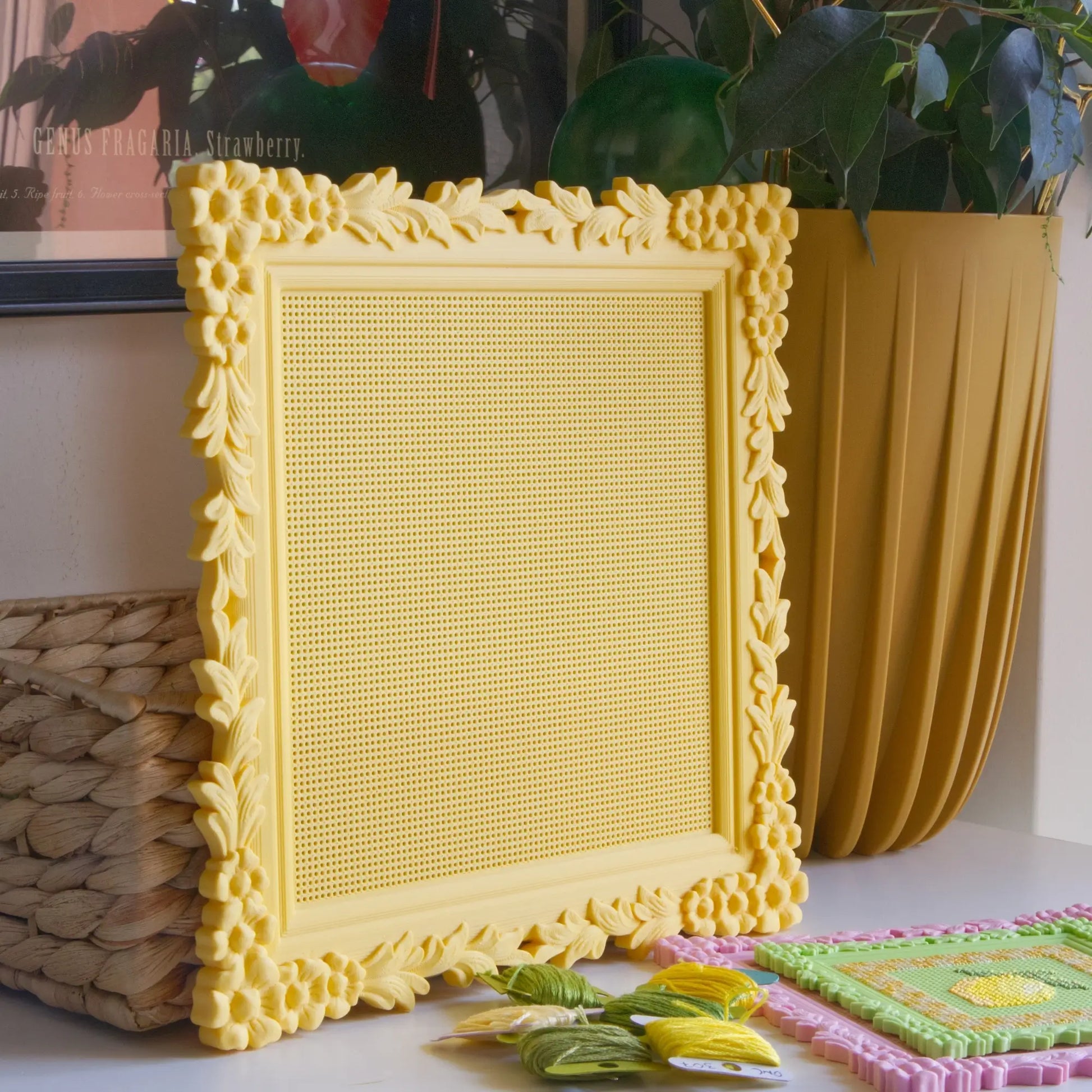 Yellow floral cross stitch frame with the blank stitchable grid, styled on a shelf with a woven basket and embroidery floss.