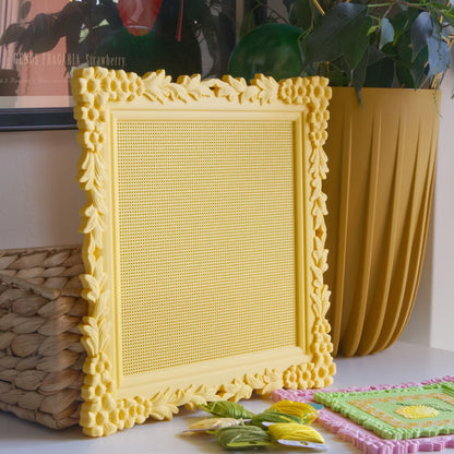 Yellow floral cross stitch frame with the blank stitchable grid, styled on a shelf with a woven basket and embroidery floss.