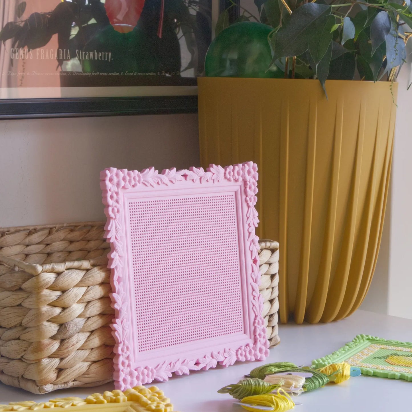 Pink floral cross stitch frame with the blank stitchable grid, styled on a shelf with a woven basket and embroidery floss.