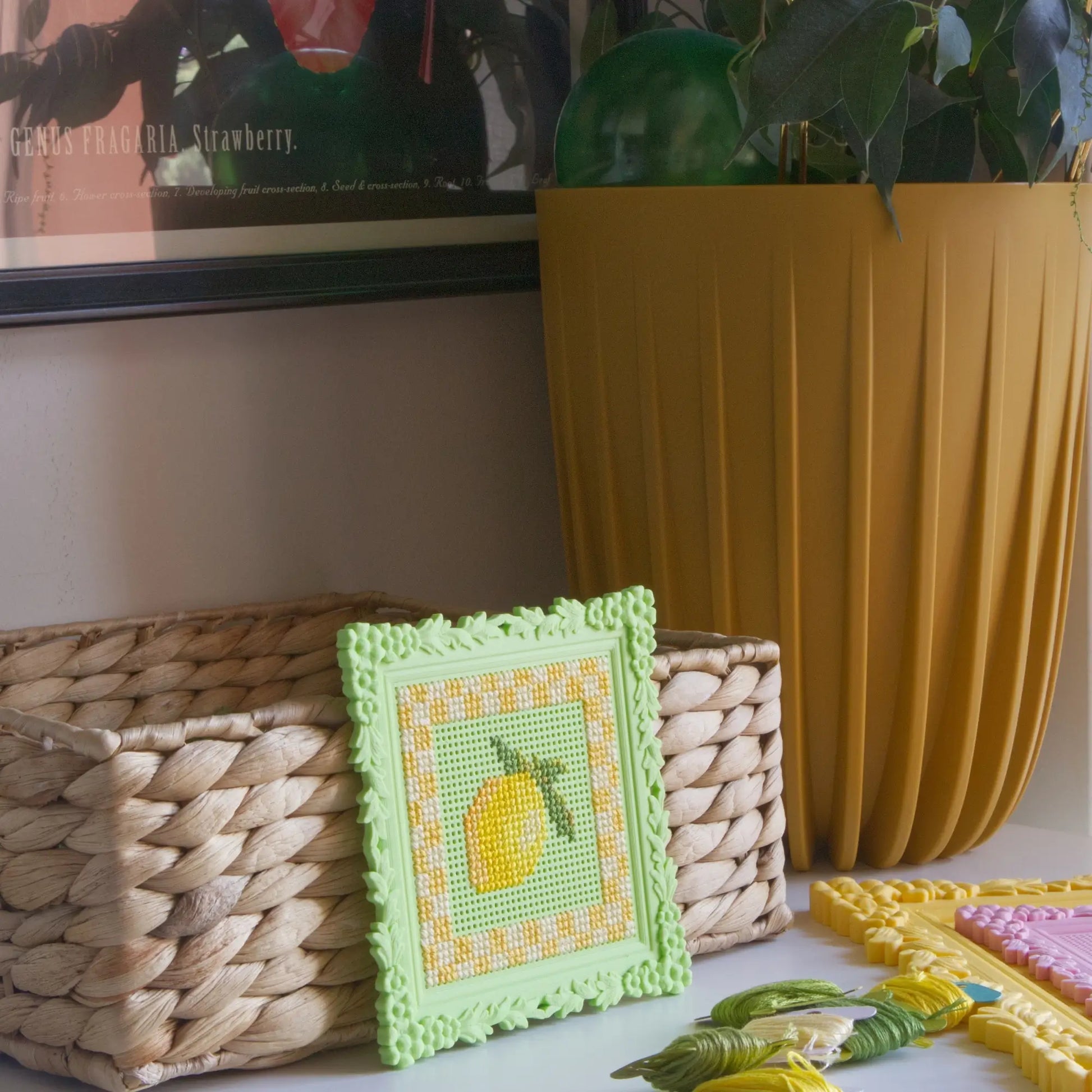 Green floral cross stitch frame with a finished lemon design, styled on a shelf with a woven basket and embroidery floss.