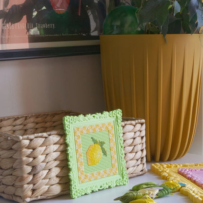 Green floral cross stitch frame with a finished lemon design, styled on a shelf with a woven basket and embroidery floss.