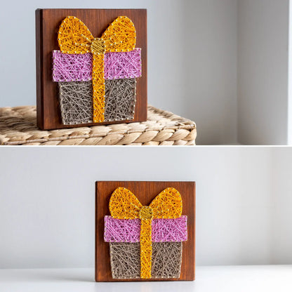 String art of a gift box with a bow on a wooden board, displayed against a neutral background.