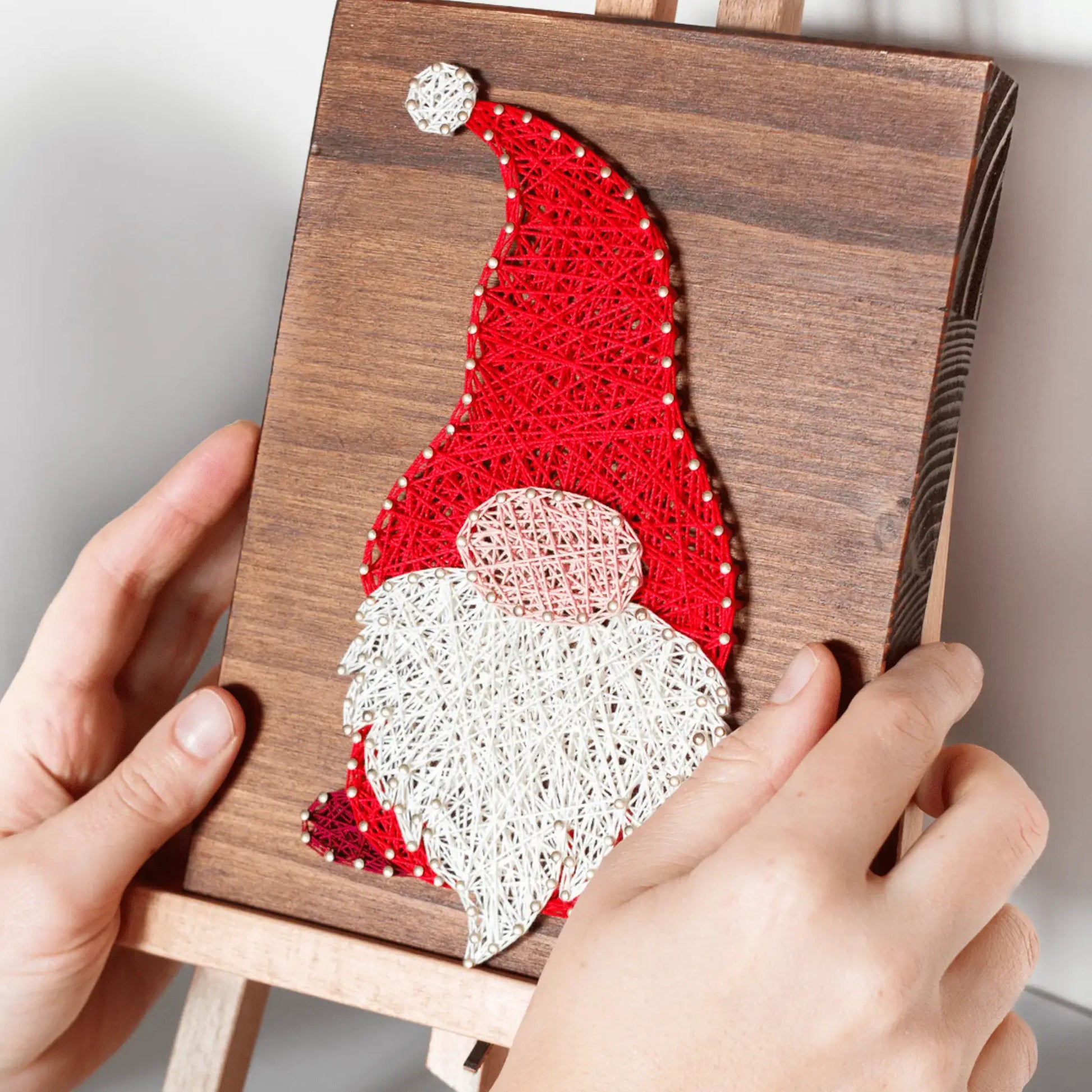 Hands holding a wooden board with completed gnome string art, showcasing a red hat and white beard design, perfect for holiday decor.