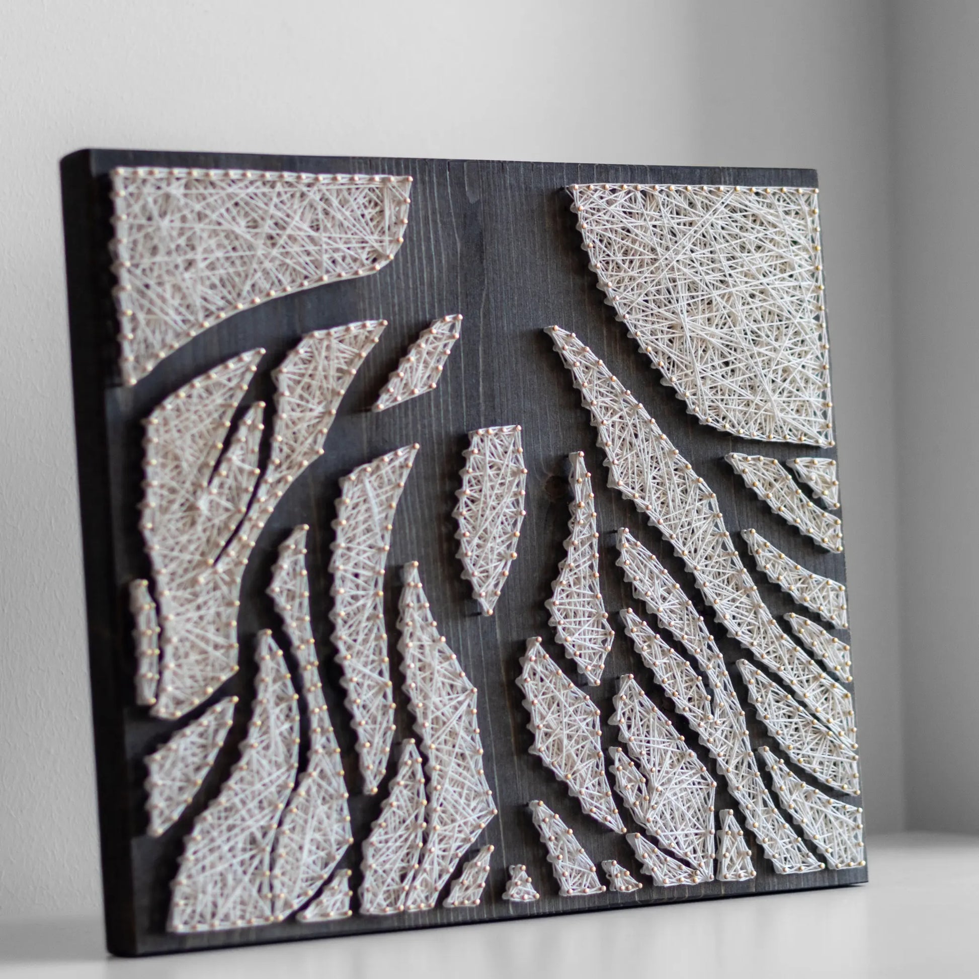 A piece of string art on a dark wooden board depicting abstract tree roots. The roots are created using white string and gold pins, arranged in various geometric shapes to form the intricate pattern of the tree roots.