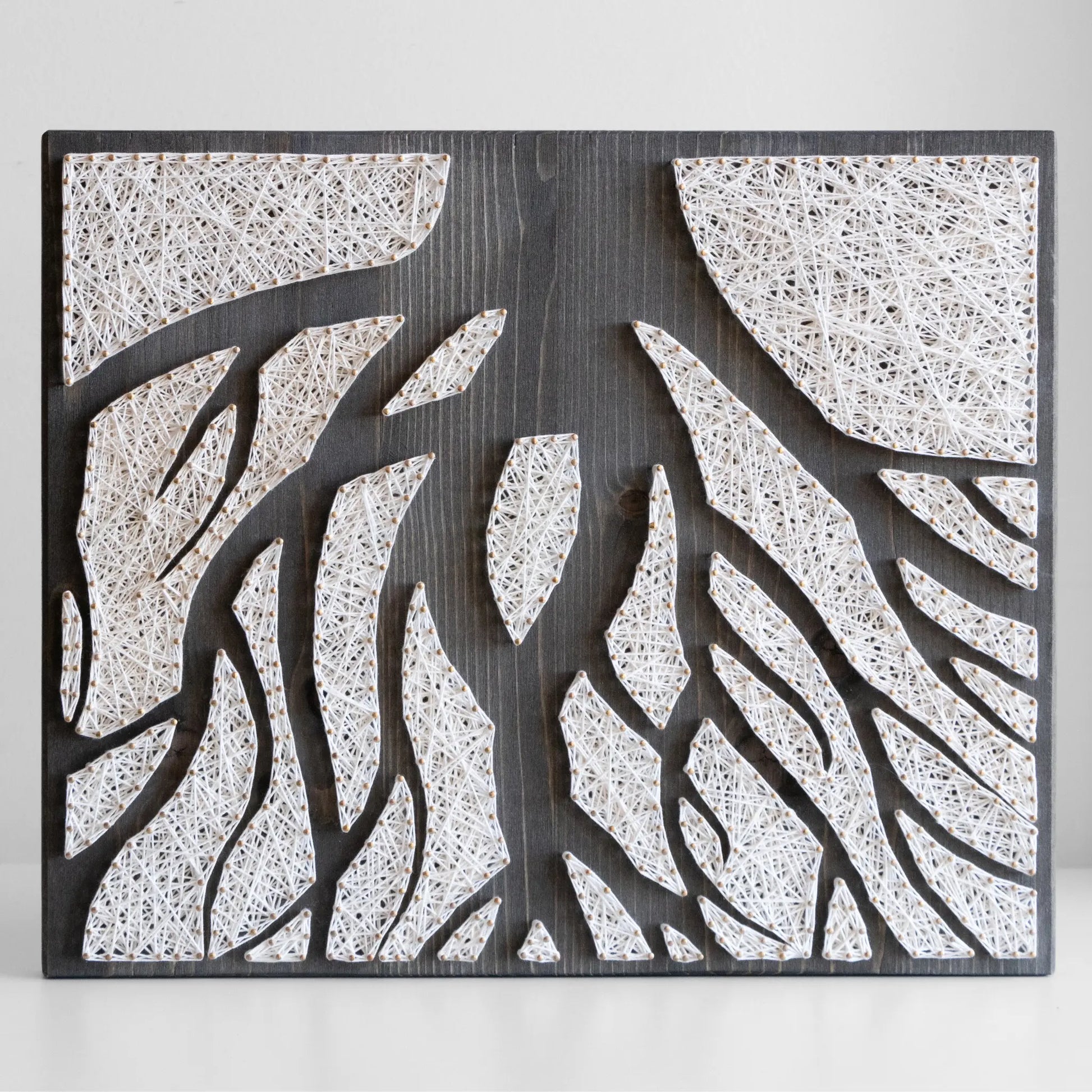 A piece of string art on a dark wooden board depicting abstract tree roots. The roots are created using white string and gold pins, arranged in various geometric shapes to form the intricate pattern of the tree roots.