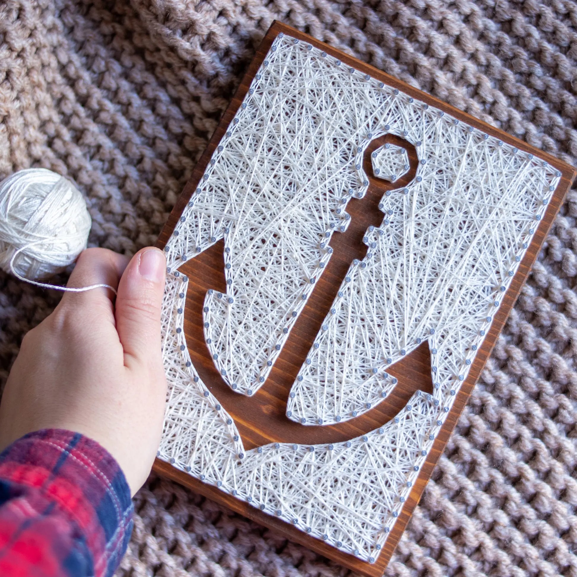 DIY string art kit featuring an anchor outlined with white thread on a rustic wooden board by GoodStrings - top view, hand holding