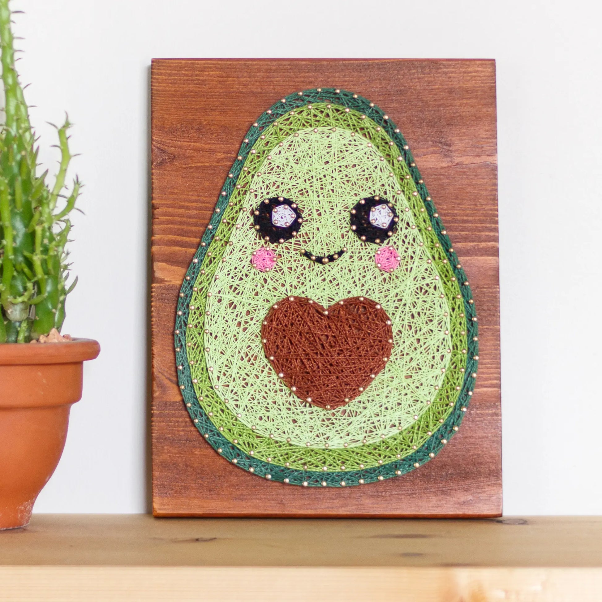 DIY string art kit featuring a cute avocado with a brown heart pit and smiling face created with threads on a wooden board by GoodStrings