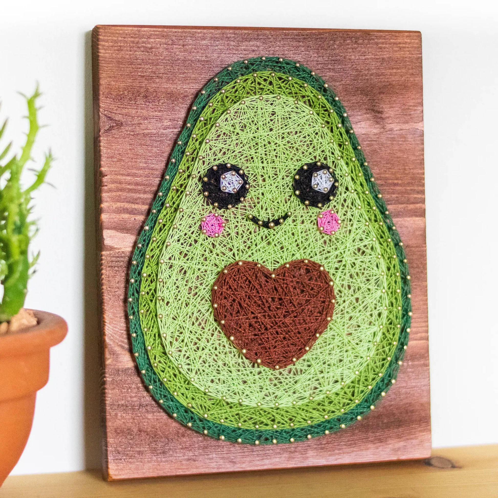 DIY string art kit featuring a cute avocado with a brown heart pit and smiling face created with threads on a wooden board by GoodStrings