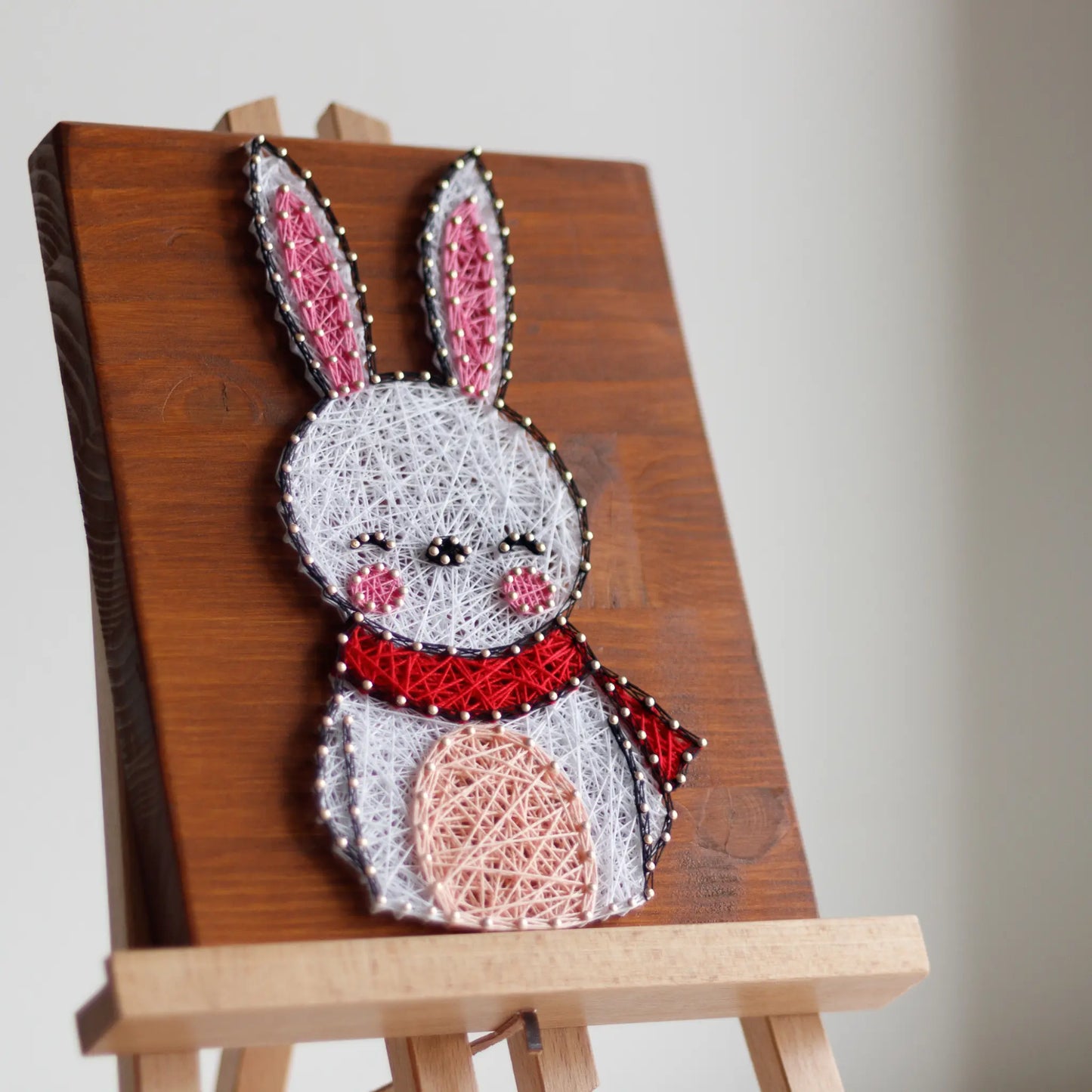 DIY string art kit featuring a cute bunny outlined with white, pink, and red thread on a rustic wooden board by GoodStrings - side view